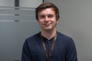 En person med ID-bånd rundt halsen står innendørs foran en vegg med matterte glasspaneler. Personen ser mot kamera og smiler svakt.