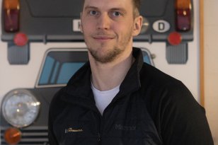 En mann i svart jakke og hvit t-skjorte. Bak han er det bilder av gamle bilder. Han er inne.