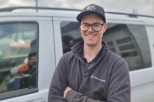 En smilende mann med briller og svart caps står med armene i kors foran en grå varebil. Han har mørk arbeidsgenser med firmalogo og ser rett mot kamera. Bakgrunnen er lett uskarp.