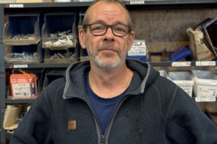 En mann med kort grått hår, skjegg og briller står foran hyller med bokser, sprayflasker og ulike deler. Han har på seg en mørk blå fleecejakke over en blå T-skjorte og ser mot kameraet.