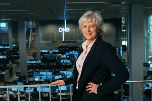 Dame med lys skjorte og svart blazer og grått hår poserer ved et rekkverk. Logoen til DNB vises i bakgrunnen. Det er også mange pc-skjermer i bakgrunnen.