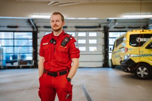 En mann med kort hår og skjegg står i en rød ambulanse-uniform inne i en vedlikeholdshall og ser på kamera. I bakgrunnen står to ambulanser.