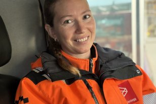 En smilende kvinne med håret i flette sitter inne i et kjøretøy og ser mot kameraet. Hun har på seg en oransje arbeidsjakke med refleksstriper og svart krage.