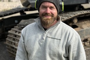 Vi ser en smilende mann med kort hår og skjegg foran en stor gravemaskin. Han har på seg en lysegrå genser, svart lue og hørselsvern. Gravemaskinens belter er synlige i bakgrunnen.