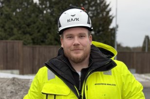 En mann med brunt skjegg, iført vernehjelm og gul refleksjakke står på en byggeplass. Han ser rett mot kamera med byggearbeid og natur i bakgrunnen.