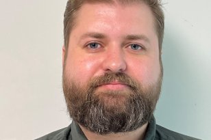 En mann med kort, lysebrunt hår og skjegg ser rett mot kamera foran en lys vegg. Han har på seg en mørkgrønn skjorte. Bildet er et tett utsnitt av ansikt og overkropp og fremstår som et portrett tatt innendørs.