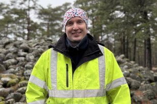 En person i gul refleksjakke og mønstret lue står ute i skogen foran en stor samling steiner. Personen smiler mot kamera. Rundt han er det furutrær og mosekledd terreng.