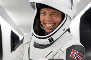 En person i hvit romdrakt smiler bredt og blunker til kamera. Hun peker på et norsk flaggemblem på drakten, hvor det også står navnet «J. Mikkelsen».