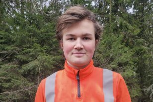 En ung mann med brunt hår smiler. Bak han er det skog. Han har på seg en oransj genser med refleksstriper. 