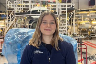 Dame med blå genser og skulderlangt hår står inne i en stor verkstedhall