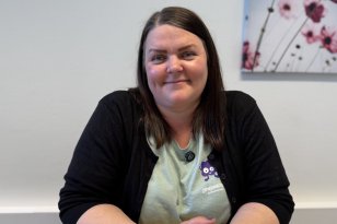 En kvinne med brunt halvlangt hår sitter ved et bord og ser mot kamera. Hun har på seg en lys grønn t-skjorte og en svart cardigan. Hun smiler og har hendene foldet foran seg på bordet. På veggen bak henne henger det et bilde av noen rosa blomster.