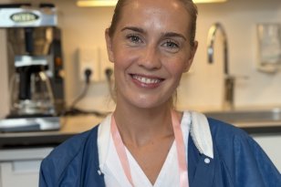 En smilende kvinne med blondt hår samlet bakover ser mot kameraet. Hun har på seg hvitt arbeidstøy med en blå frakk over og et rosa bånd med ID-kort rundt halsen. I bakgrunnen ses et kjøkken- eller pauserom med blant annet kaffetrakter, skap og vask.