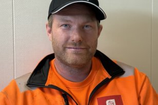 En mann med kort skjegg og blå øyne ser mot kameraet. Han har på seg en svart caps med hvite bokstaver, oransje arbeidsjakke med refleks og en oransje T-skjorte.