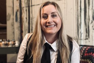 En dame med langt blondt hår smiler. Hun har på seg hvit skjorte, med svart vest og svart slips. Hun sitter på en brun stol og det ligger et lappeteppe bak hun. I bakgrunnen er det maling og en vegg som har blitt malt på.