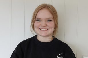 Jente med blondt hår i hestehale som smiler til kamera. Hun har på svart genser med hvit logo.