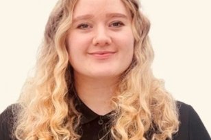 Portrett av en ung kvinne med lyst, krøllete hår som faller ned over skuldrene. Hun ser rett i kamera og smiler svakt. Hun har på seg en svart skjorte med krage og en liten logo på brystet. Bakgrunnen er lys og nøytral.