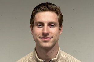 Mann med brunt hår og beige genser står foran en beige vegg og smiler til kamera