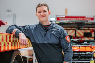 En mann i brannuniform står lent opp mot en brannbil mens han smiler til kamera.