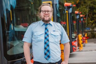En mann med langt hår og skjegg står foran en rad med busser og smiler til kamera. Han har på uniform med lyseblå skjorte, slips og mørk bukse. I beltet er det festet et nøkkelknippe.
