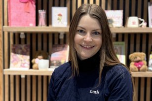 Dame med brunt hår og blå genser med logo står bak en disk med en scanner i den ene hånda. I den andre hånda har hun en bok.