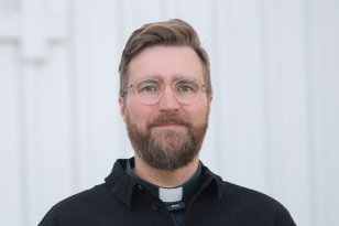 Mann med blondt kort hår, skjegg og briller ser rett frem. Han har på svart skjorte og en mørk grønn skjorte under. Han bærer en hvit snipp i halskragen. 