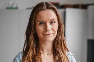 En kvinne med mørkt hår smiler til kamera. Hun har på seg en hvit singlet under en lyseblå skjorte, og et smykke rundt halsen.