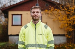 En mann i refleksjakke står foran et eldre, lite bygg og smiler til kamera. Han har hendene i lommene, og logoen til Inotek på brystet.