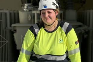 En kvinne med hvit vernehjelm og gul arbeidsgenser med refleks sitter foran et stort elektrisk anlegg. Hun smiler svakt mot kameraet. I bakgrunnen er det store grå transformatorer og annet elektrisk utstyr.