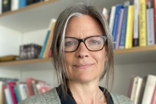 Dame med briller og svart t-skjorte og grå cardigan ser inn i kamera. I bakgrunnen er det tre bokhyller på en hvit vegg.