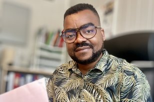 Flyktningkonsulent Akili Christophe Kahindo på kontoret sitt. I hånden har han en blyant og ark. Mann med mønstrete skjorte og briller sitter ved en kontorpult og skriver på et ark med en blyant