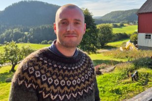 En mann med kort hår og mørk, mønstret strikkegenser står utendørs og ser mot kameraet. I bakgrunnen er det grønne jorder, skogkledde åser og en rød gårdsbygning.
