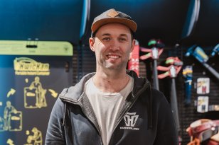 En mann med hettejakke og caps står foran en utstillingsvegg i en butikk, full av verktøy. Han har hendene i lommene og smiler til kamera.