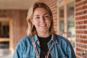 En kvinne med blondt hår, en stor blå skjorte og et nøkkelbånd rundt halsen smiler til kamera. I bakgrunnen er en skolebygning med en rødlig teglsteinsvegg.