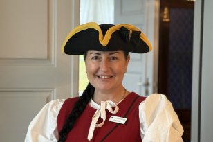 En smilende kvinne med langt, mørkt hår i flette står i en døråpning. Hun har på seg en rød vest over en hvit bluse med puffermer og en svart, bredbremmet hatt med gul kant. I bakgrunnen ses et rom med blåmønstret tapet og en lysekrone.