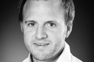 Mann med hvit skjorte ser inn i kamera. Han står foran en mørkegrå vegg.