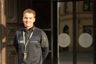 Ung mann i sort genser med logoen til Drammen Kommune på brystet står utenfor skyvedører og smiler. 