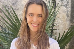 En dame med brunt hår og hvit skjorte smiler mens håret flagrer. Bak hun er det et grønt palmetre og murvegg.