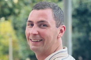 En mann smiler mot kamera i utendørs omgivelser med grønne trær i bakgrunnen. Han har kort, mørkt hår og bruker en lys skjorte med smale striper. Bildet har en varm og naturlig tone, med et avslappet uttrykk.