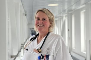 Indremedisiner Anna Sypula Nordlie En kvinne i hvit uniform står i en lys sykehuskorridor og smiler mot kamera. Hun har stetoskop rundt halsen og flere penner i brystlommen, sammen med et ID-kort som henger fra lommekanten.