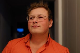 En mann står innendørs i en døråpning, iført en oransje skjorte med åpen krage. Han har kort hår og bruker runde briller. Personen ser litt opp og til siden. Bakgrunnen er dempet og mørk med svakt lys som kommer fra siden.