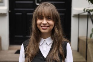 Bildet viser en ung kvinne som står utendørs foran en bygård med mørk inngangsdør. Hun smiler og ser rett mot kameraet. Hun har langt, lysebrunt hår med pannelugg, og hun er kledd i en hvit skjorte med en sort vest utenpå. Bak henne ser man steintrapper og litt grønt løv fra en plante ved siden av trappen.