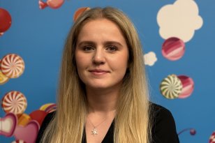 En ung kvinne med langt blondt hår står vendt mot kamera foran en blå vegg dekorert med illustrasjoner av godteri og skyer. Hun har på seg en sort topp og et smykke rundt halsen.