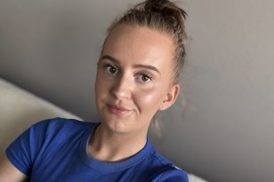 En kvinne sitter i en sofa og ser inn i kamera. Hun har håret satt opp i en topp og har på seg en blå t-skjorte. Hun smiler svakt, og bildet er tatt tett på ansikt og overkropp mot en nøytral bakgrunn.