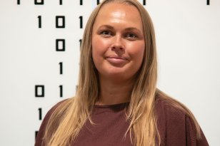 Kvinne med langt blondt hår og vinrød bluse smiler foran en vegg med binære tall.