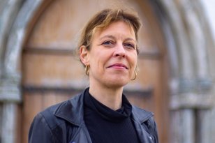Kantor Maria-Daniela Raasch Kvinne med svart genser og svart skinnjakke. Hun har runde gulløredobber og vind i håret. Hun smiler. Kvinnen står ute foran en kirkedør av tre.