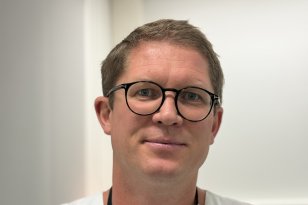 En mann med kort hår og svarte briller er fotografert i hvitt arbeidstøy. Han står foran en lys vegg og ser mot kameraet.
