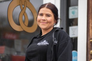 Konditor Marielle Jenssen En ung kvinne ikledd svart tskjorte og svart jakke står utenfor et bakeri og smiler til kamera. Hun har på seg uniform, med logo der det står "BLOMSØ". Bak henne henger et stort gyldent skilt av en kringle med en krone.