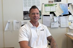 En mann med kort, mørkt hår sitter ved skrivebordet på kontoret. Han har på seg en hvit poloskjorte med TESS-logo og svarte bukser. Begge underarmene er dekket av tatoveringer. Han smiler mot kameraet, og bak ham henger blant annet kalender, oppslag og dokumenter.