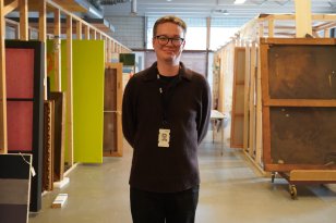 Mann med svart genser og svart bukse og briller og nøkkelkort smiler til kamera med mange lerreter i bakgrunnen