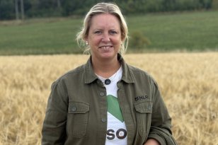 En kvinne står foran en kornåker og smiler mot kameraet. Hun har på seg en grønn jakke over en hvit T-skjorte. I bakgrunnen er det grønne jorder og skog.